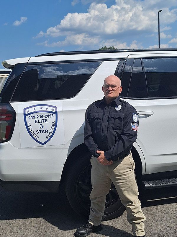 Man in security uniform stands by a white SUV with security company logo.