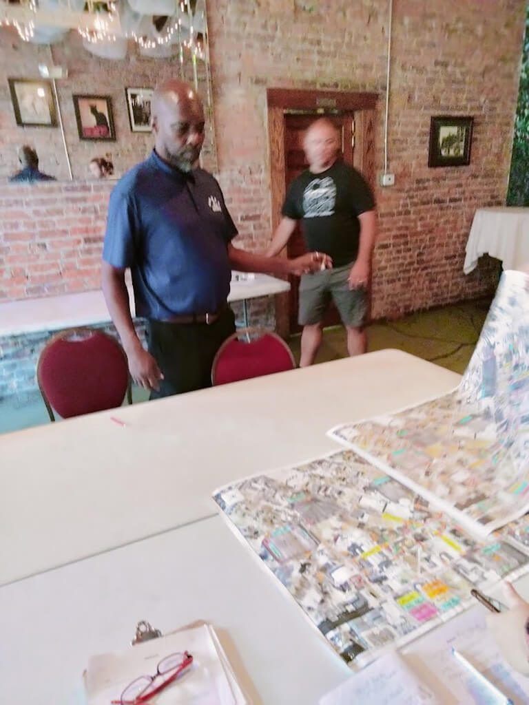 Two men at a table with maps. One man in blue shirt points. Brick wall background.