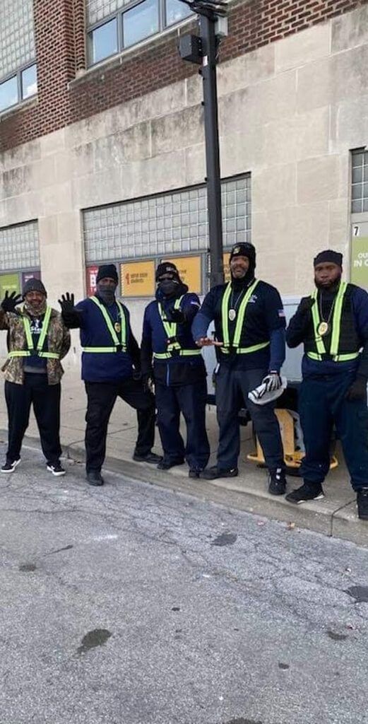 Five men in safety vests on a city sidewalk, one waves. They stand in front of a brick building.