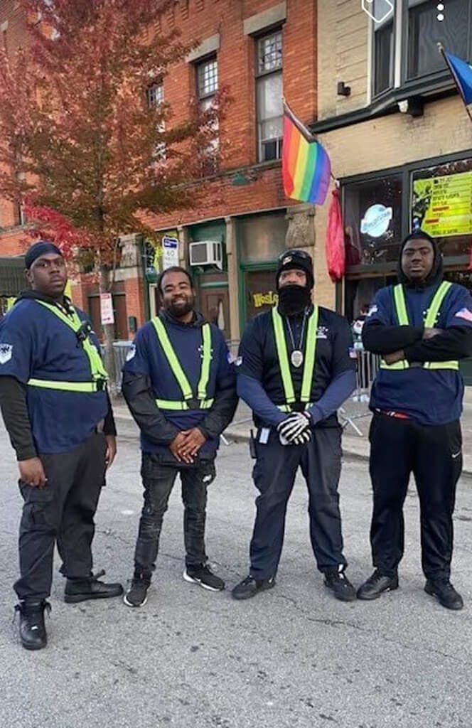 Four security guards in blue uniforms stand in front of a building with a rainbow flag.