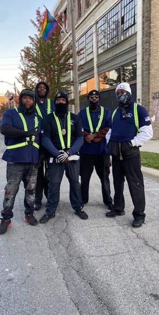 Group of men in uniforms, posing on a street. Some wear reflective vests, one holds a rainbow flag.