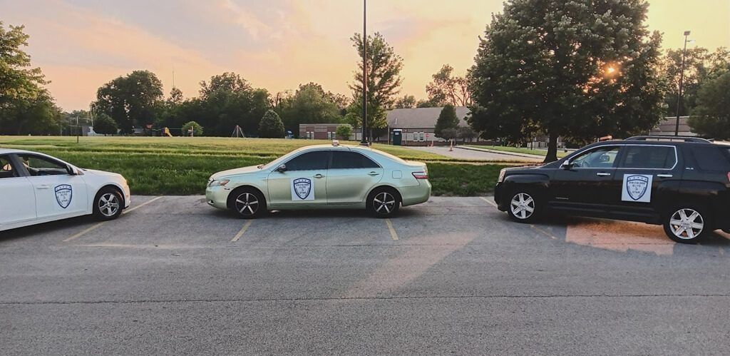 Three cars parked in a lot. White, light green, and black with a crest logo on the sides. Park lot, sunset.
