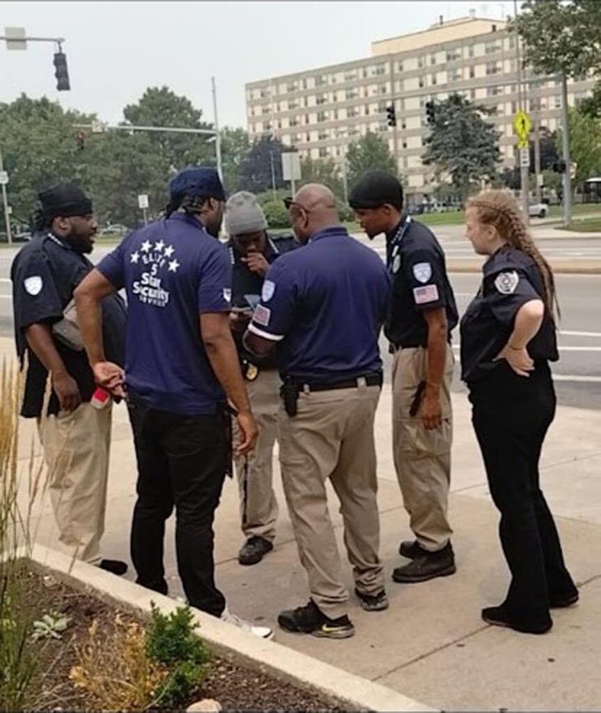 Group of security personnel in uniform looking at a phone on a sidewalk near a building.