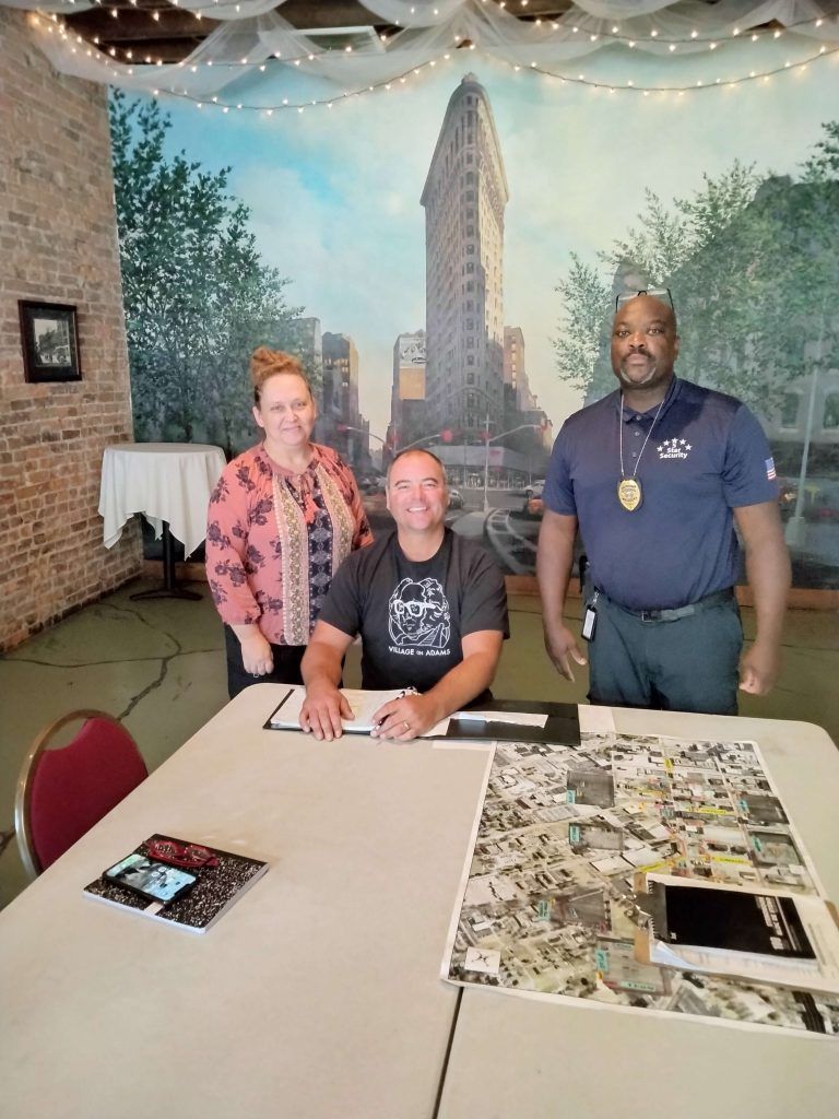 Three people at a table in front of a mural; man signing document, woman and man standing, smiling.