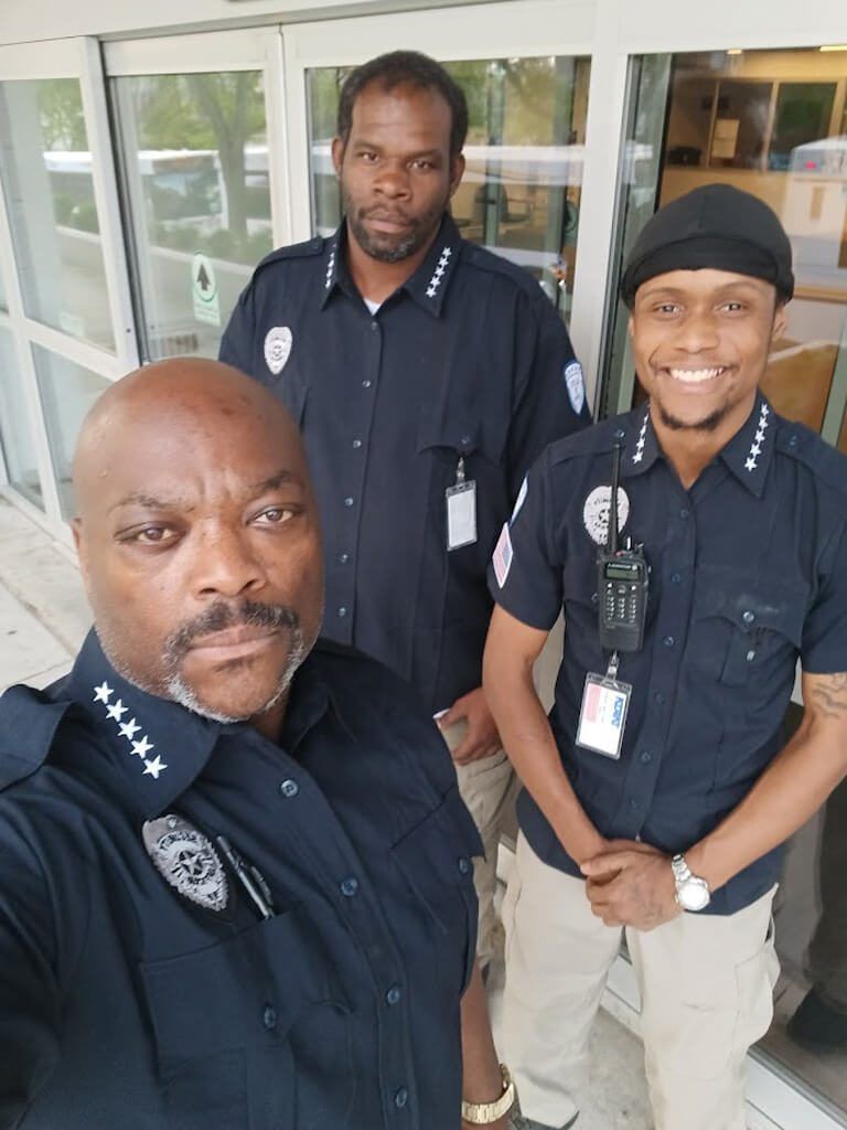Three Black men in security guard uniforms stand outside, smiling and posing.