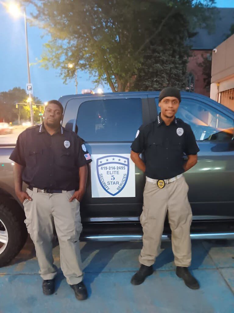 Two security guards stand next to a truck. One wears a cap. They are by a building with a logo.