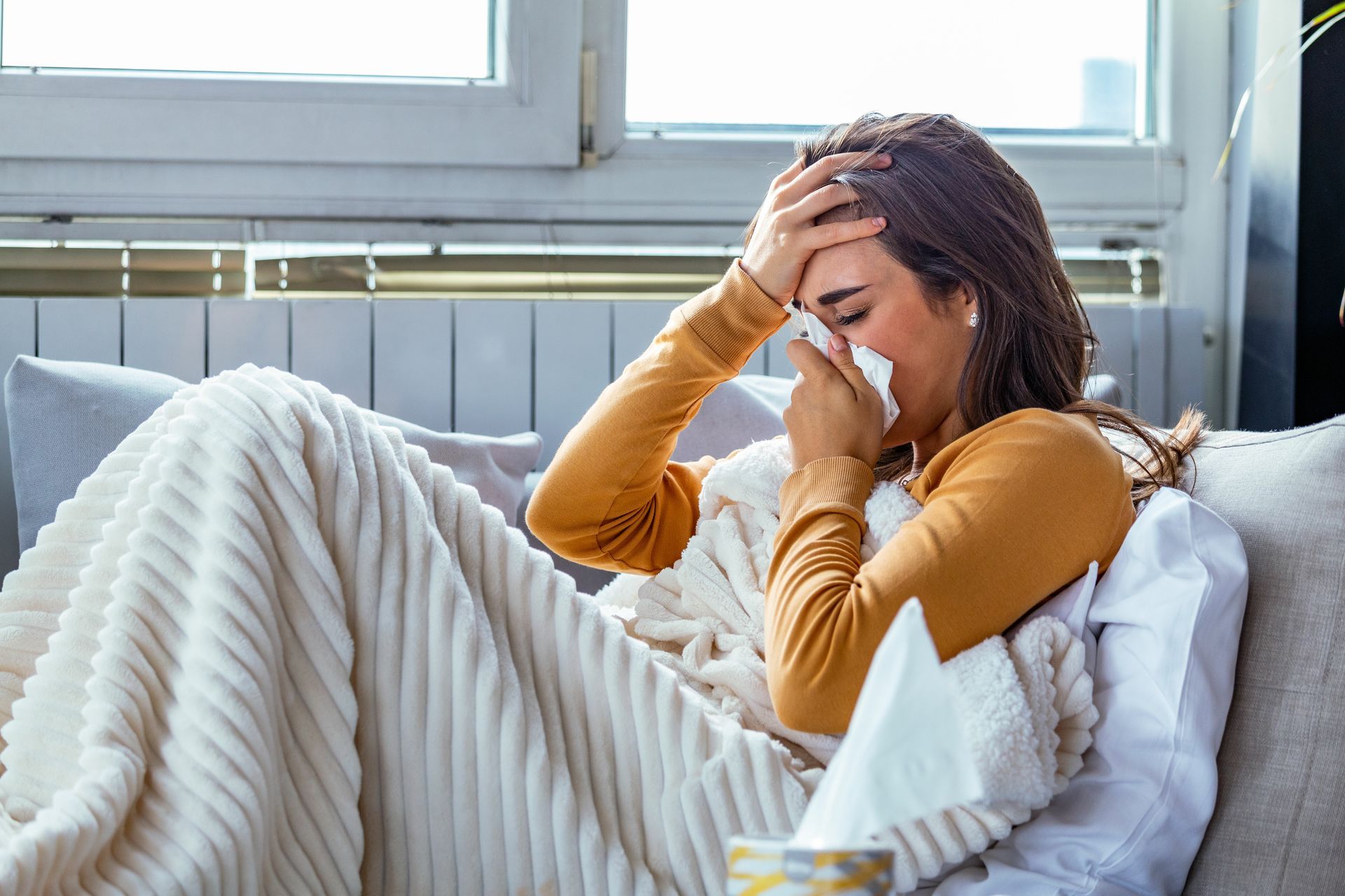 A woman is laying on a couch blowing her nose into a napkin.