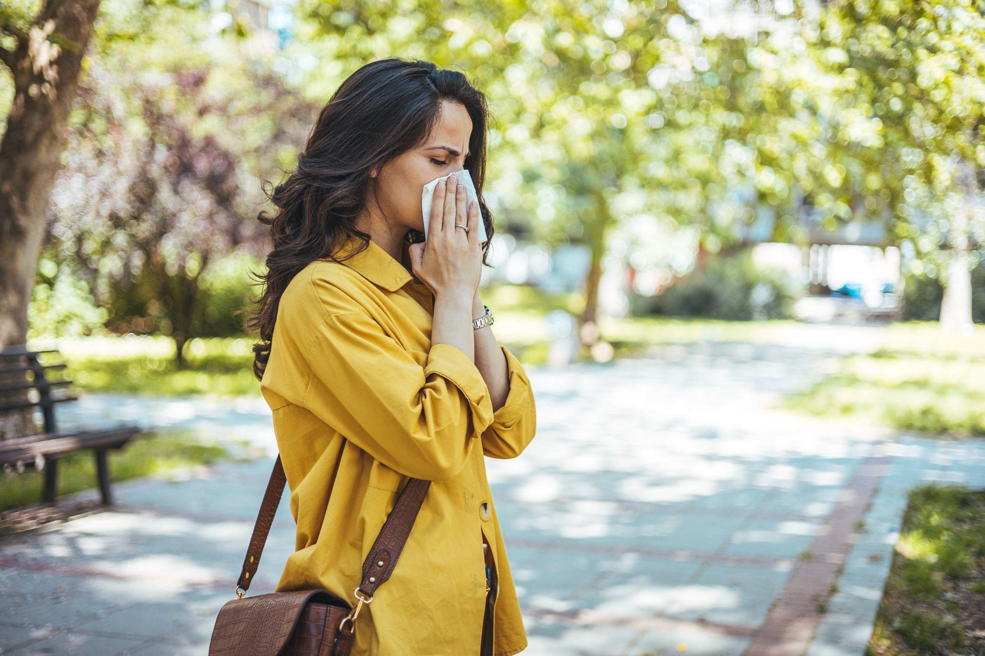 A woman in a yellow shirt is blowing her nose in a park.