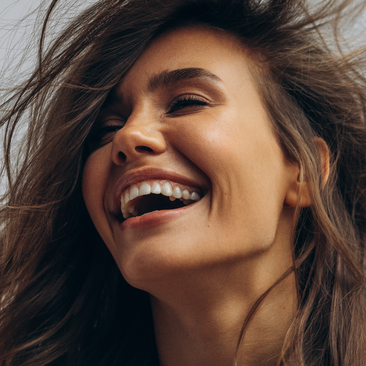 Close-up of a person with wind-swept hair, smiling broadly with their head tilted back against a soft, light background.