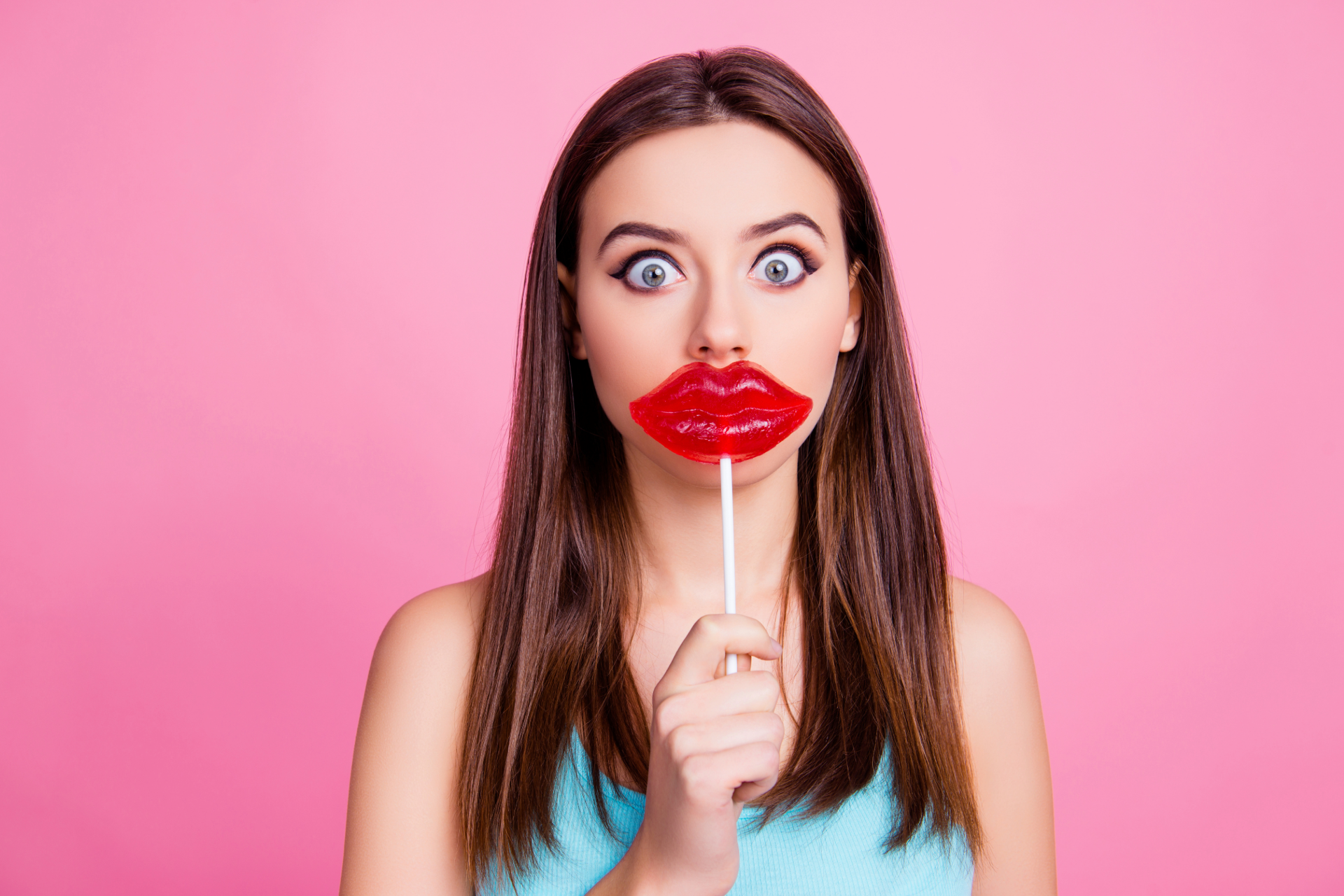 Woman with wide eyes holding a lollipop shaped like red lips in front of her mouth on a pink background.
