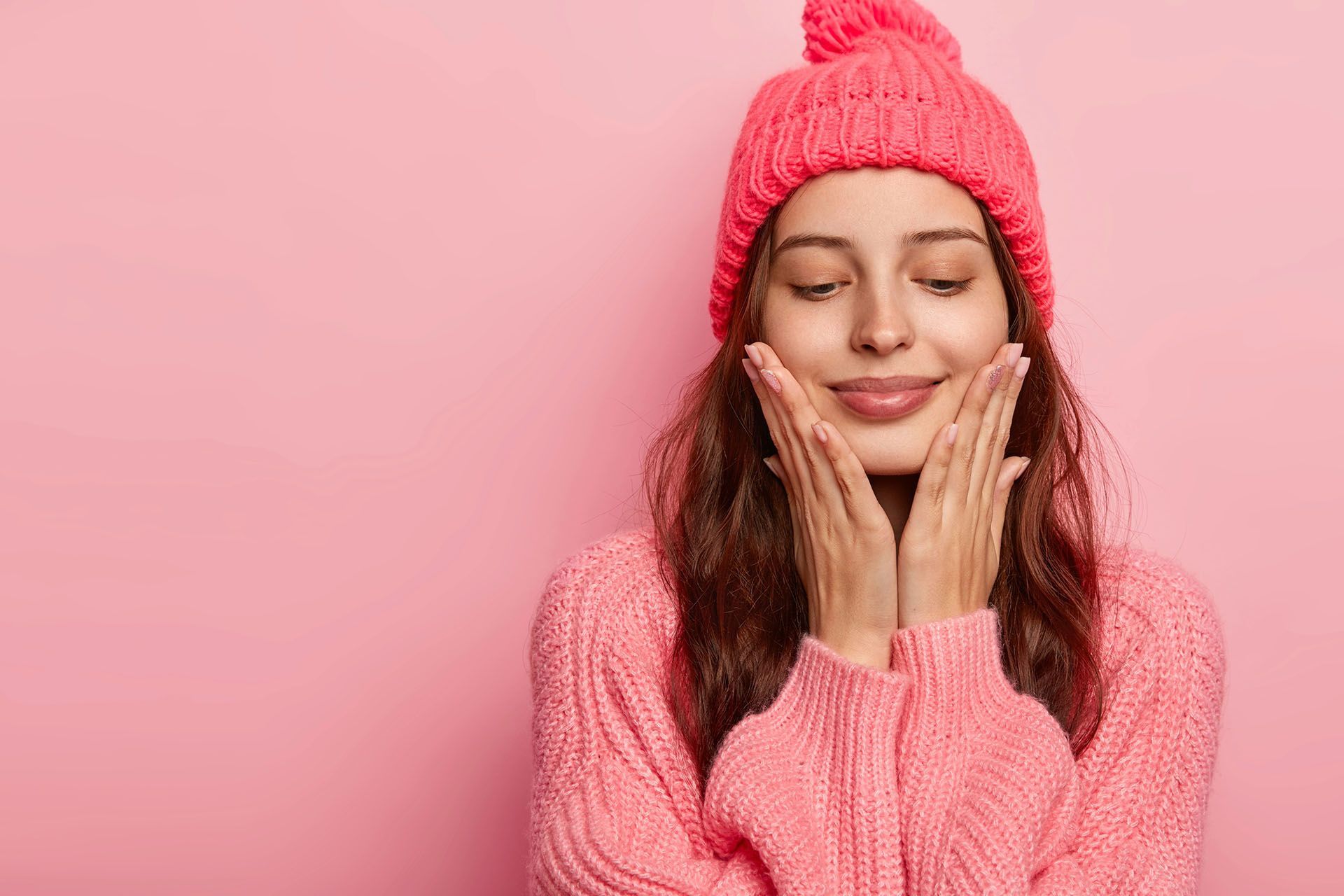 A woman in a pink hat and sweater is touching her face with her hands.