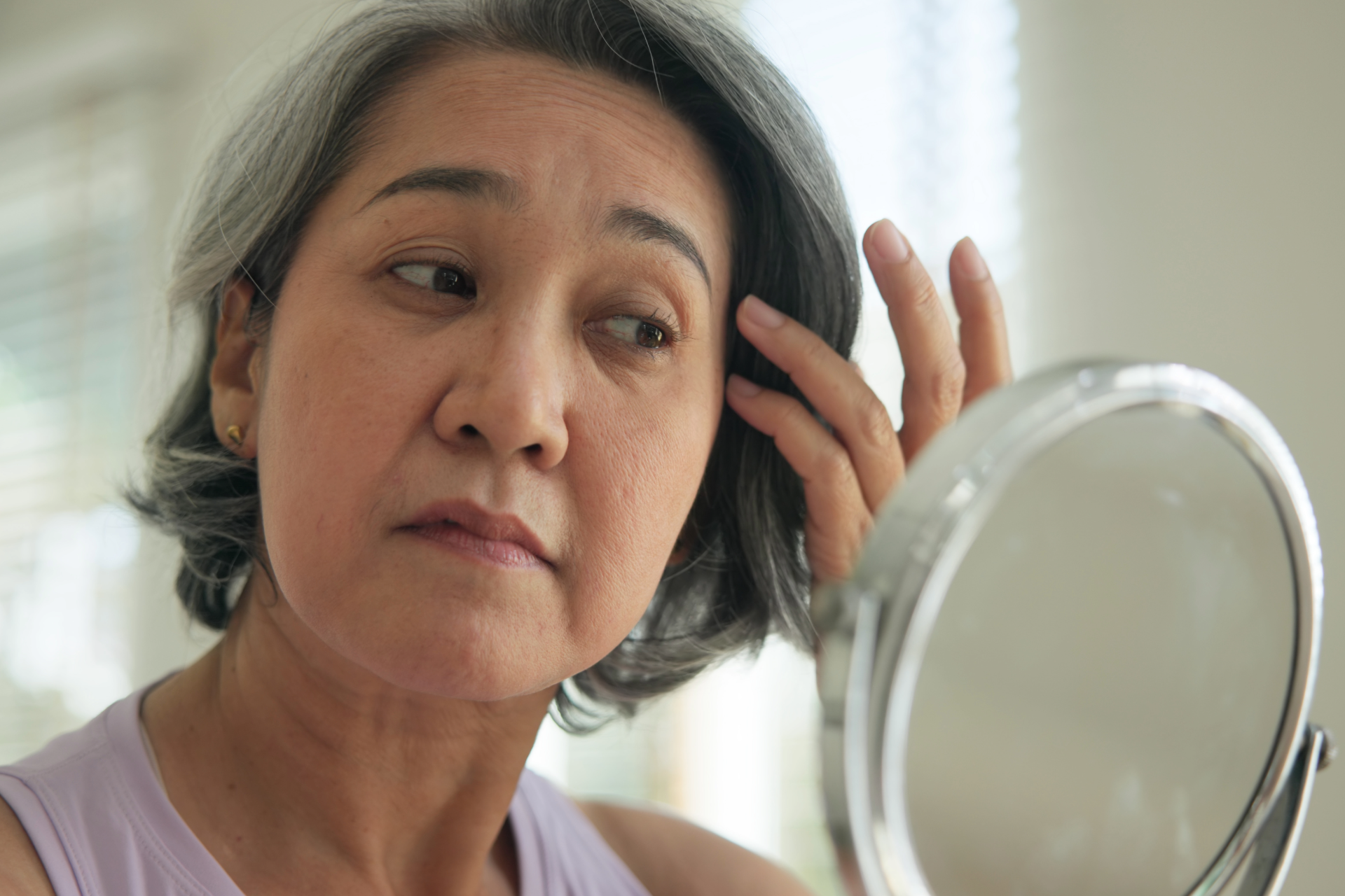 Woman with gray hair looking at a mirror, examining her face.