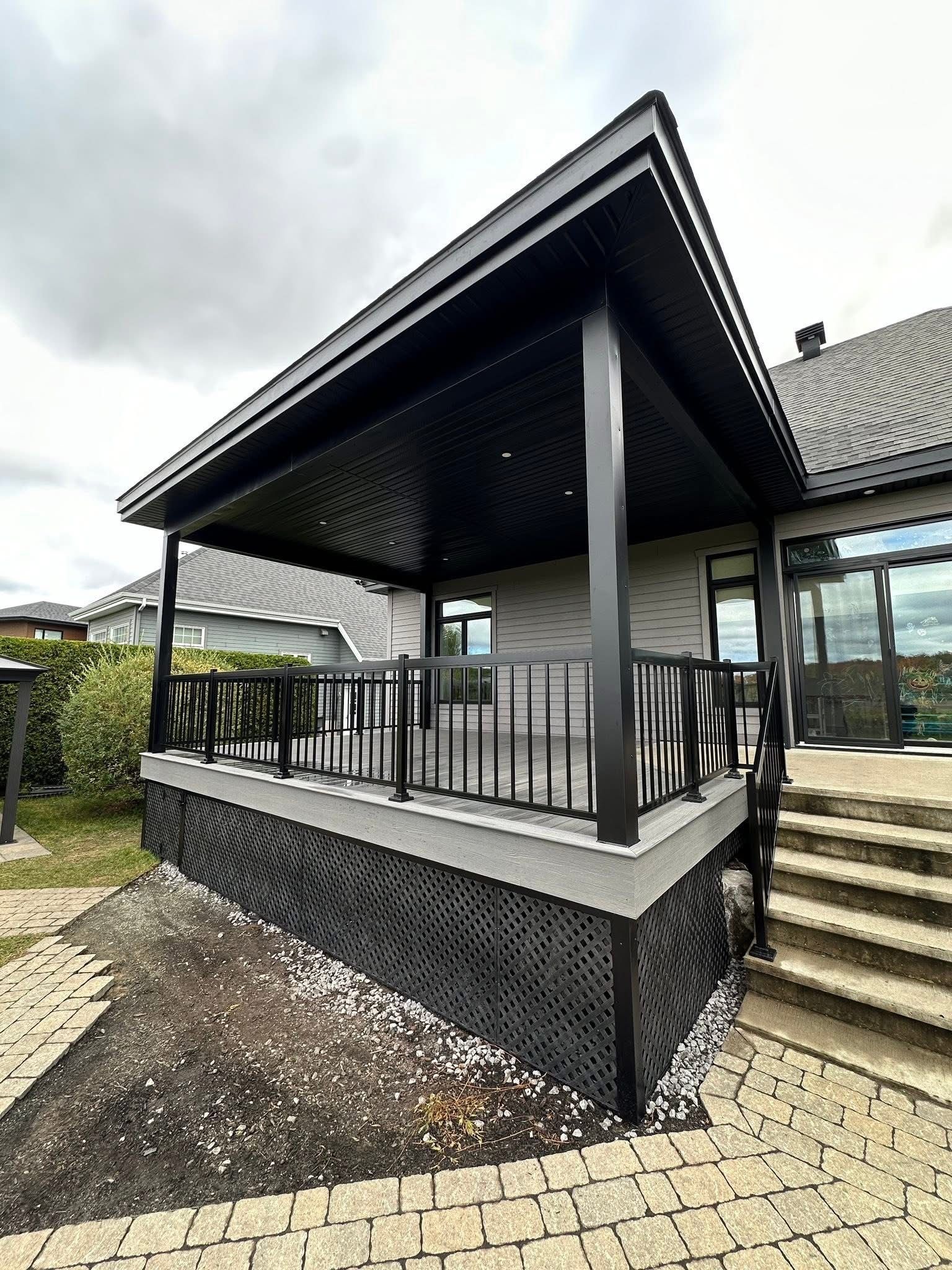 Une terrasse extérieure couverte, dotée de balustrades noires et d'une plinthe en treillis diagonal assortie, jouxte une maison au revêtement en pierre.