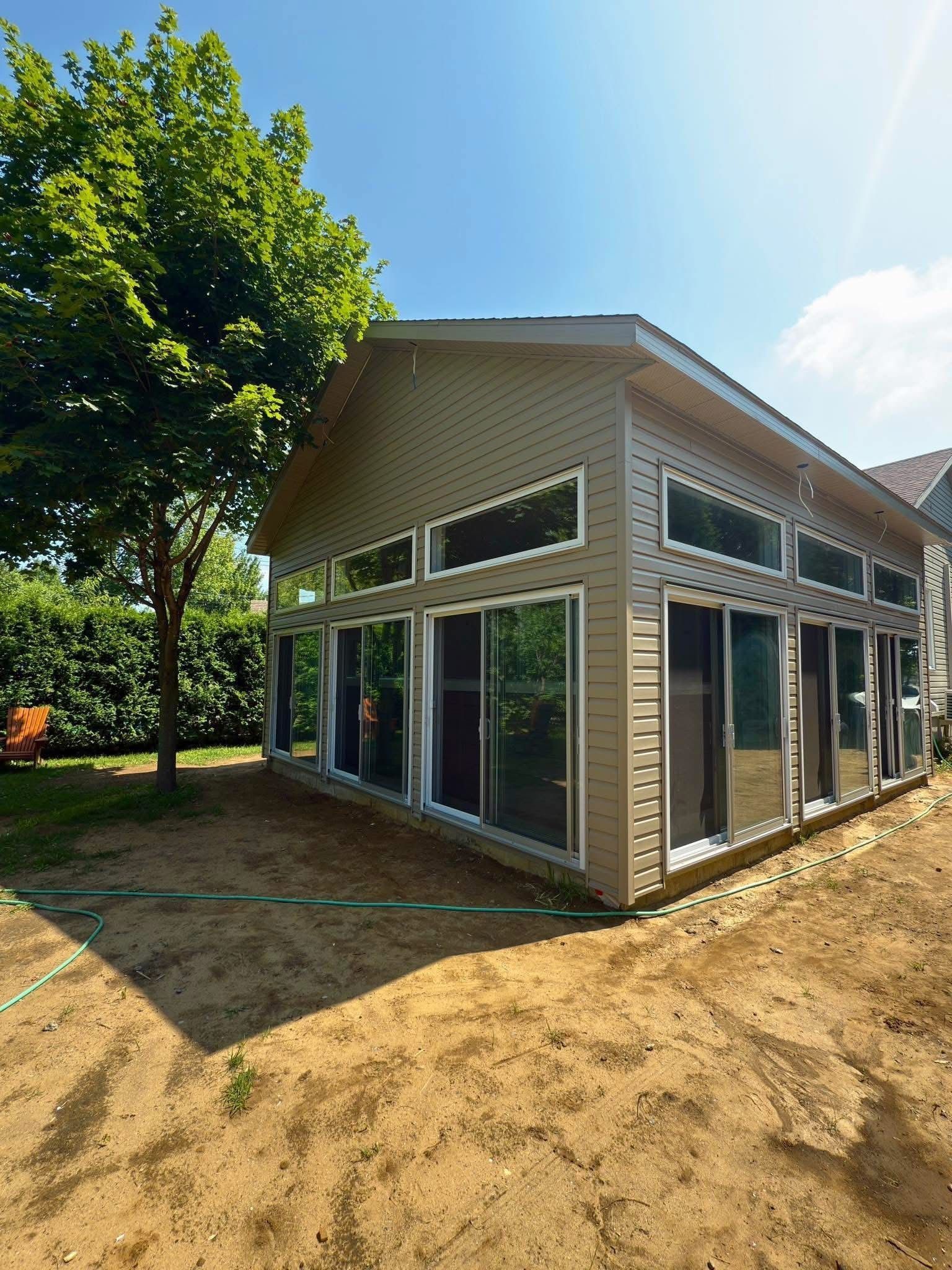 Une véranda moderne avec un revêtement extérieur en vinyle beige et de grandes fenêtres allant du sol au plafond, située dans une cour au sol nu.