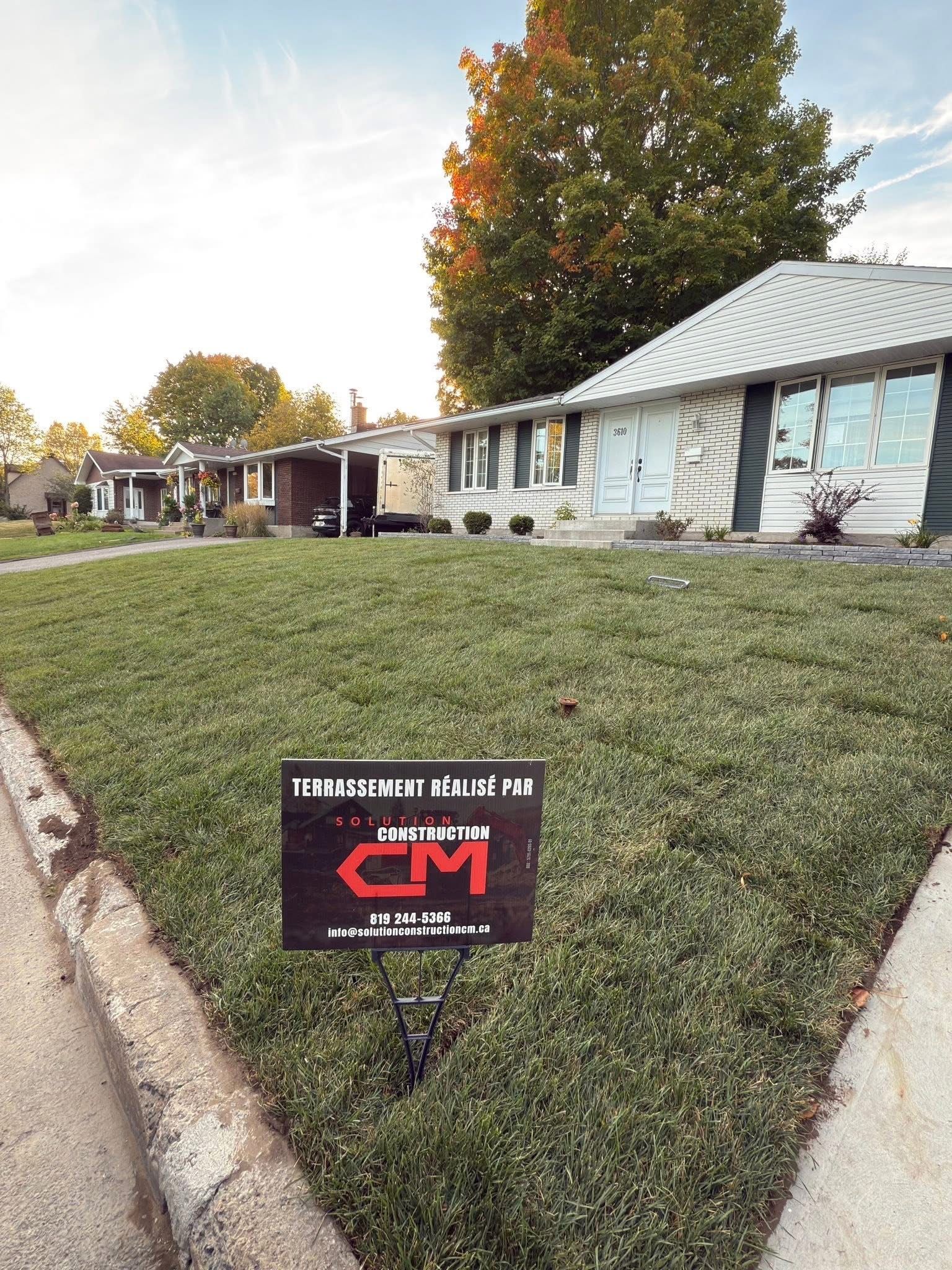 Un panneau orné du logo rouge « CM » se dresse sur une pelouse de banlieue, devant plusieurs maisons de plain-pied, sous un ciel dégagé.