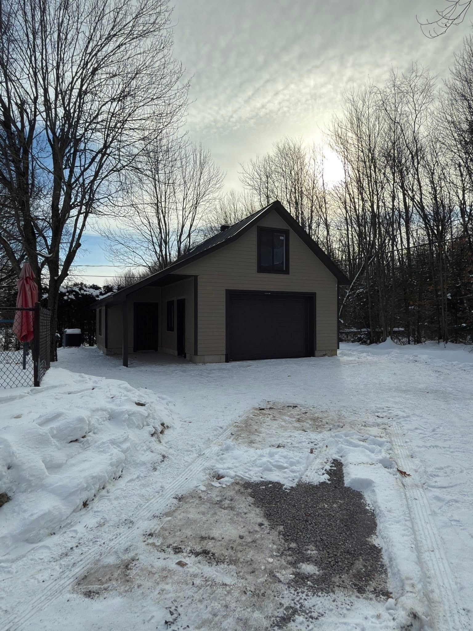 Un garage indépendant beige avec une porte et une fenêtre sombres, niché parmi les arbres et le sol enneigés par une journée d'hiver ensoleillée.