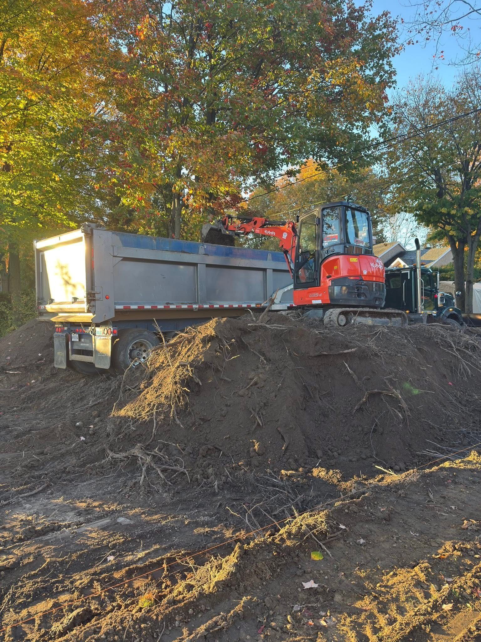 Une pelleteuse orange, sur un grand tas de terre, charge un camion-benne gris stationné près d'arbres d'automne.