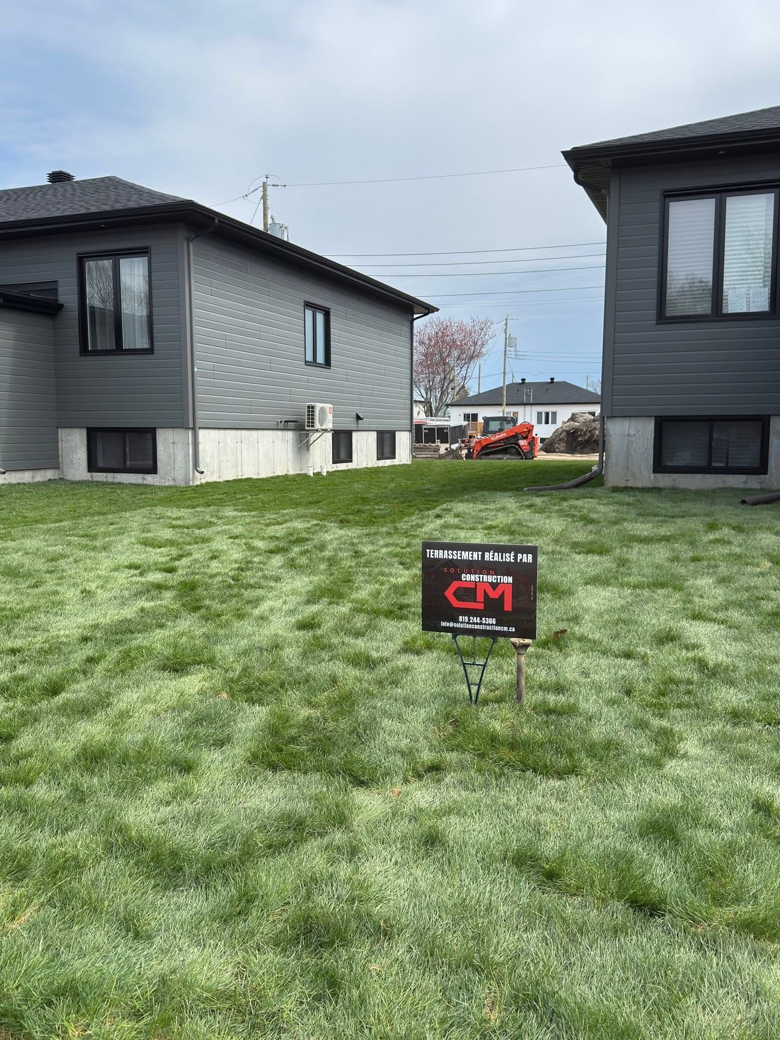 Un panneau indiquant « Maisons MD » se dresse sur une pelouse entre deux maisons grises, avec une autre maison et un chantier de construction au loin.