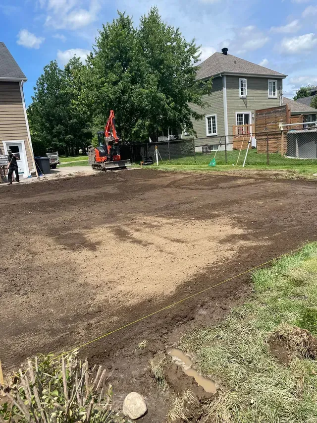 Une petite pelleteuse est en train de creuser un jardin ; de la terre est répandue. Une maison et des arbres se dressent à l’arrière-plan.
