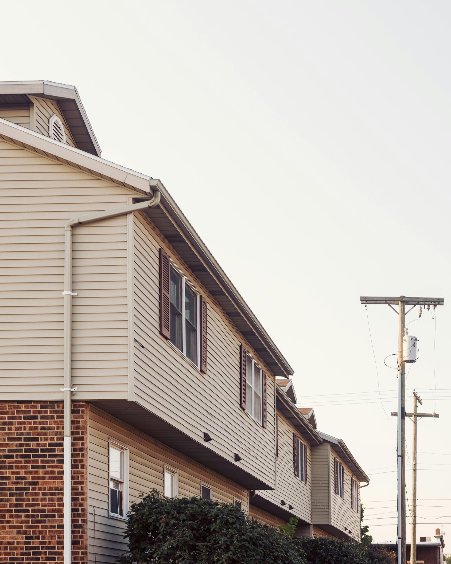 Vue en contre-plongée de maisons de ville revêtues de vinyle beige et construites sur un socle en briques, à côté d'un poteau électrique sous un ciel lumineux.