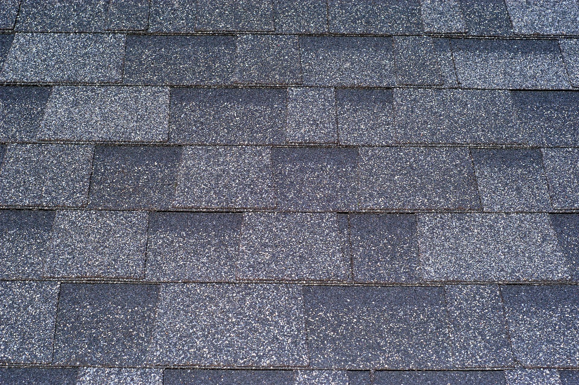 Roofer in safety vest and hardhat on a rooftop installing gray tiles against a blue sky.