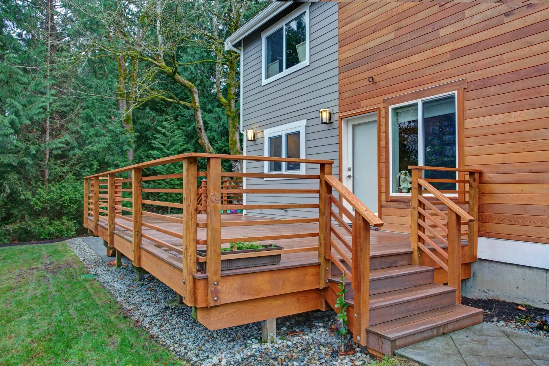 Wooden deck with stairs leading to a two-story house with cedar siding and gray siding, next to a wooded area.