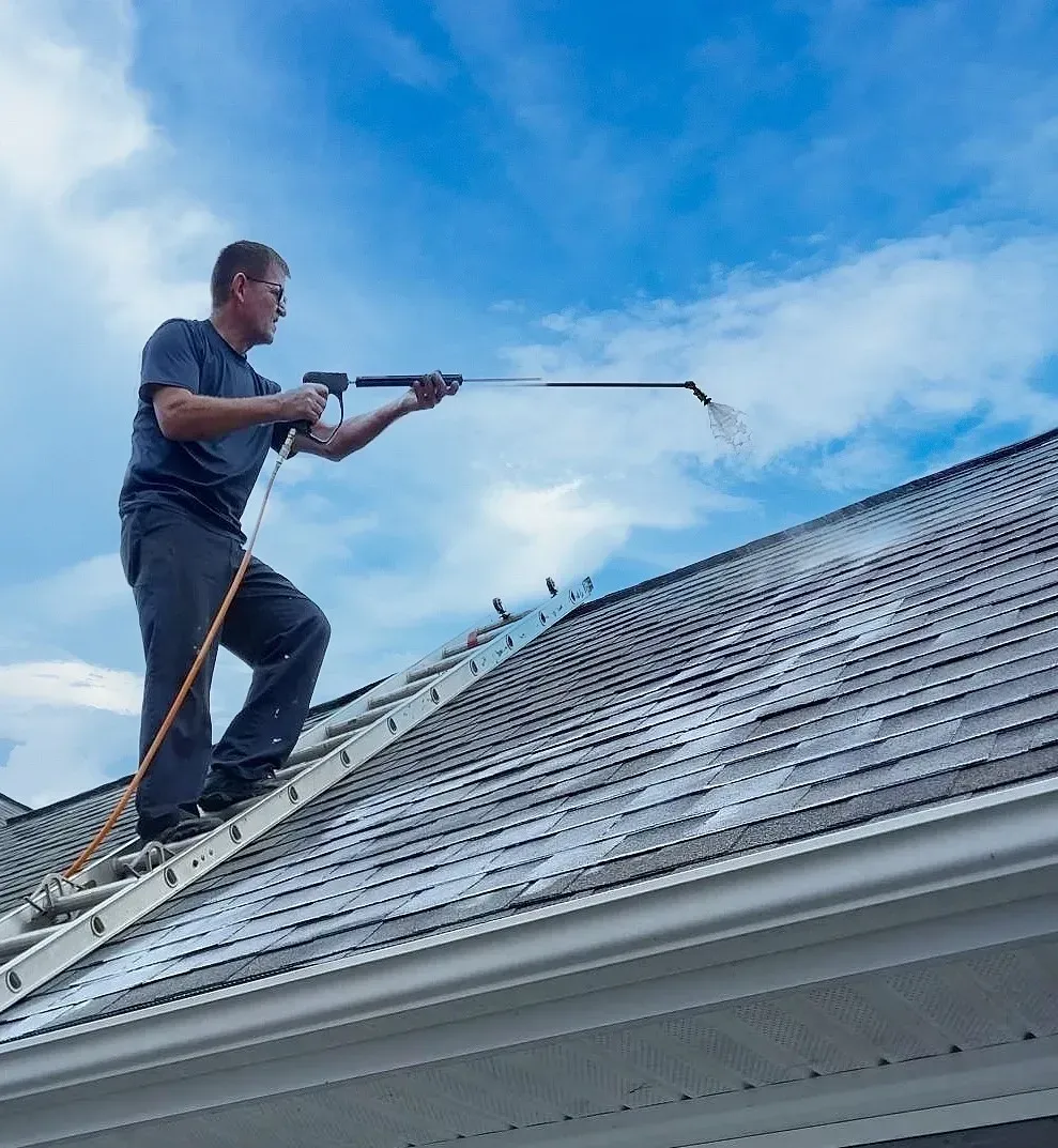 Man on ladder power washing a roof against a blue sky.