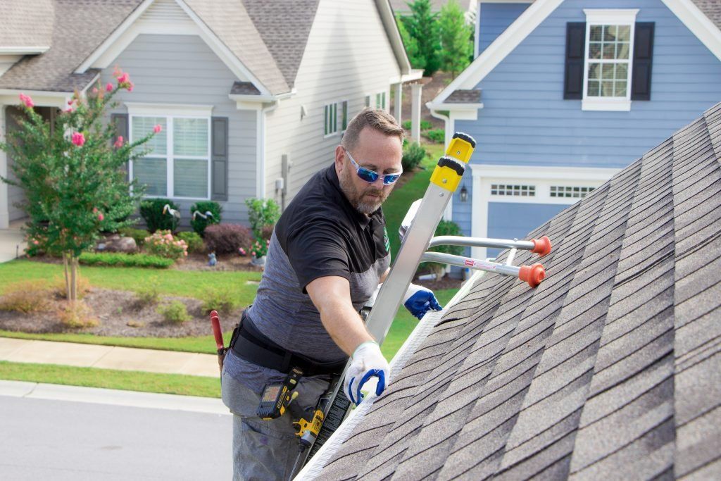 Roofer on a roof inspecting shingles, holding a ladder, wearing safety glasses, a tool belt, and gloves. Houses in background.