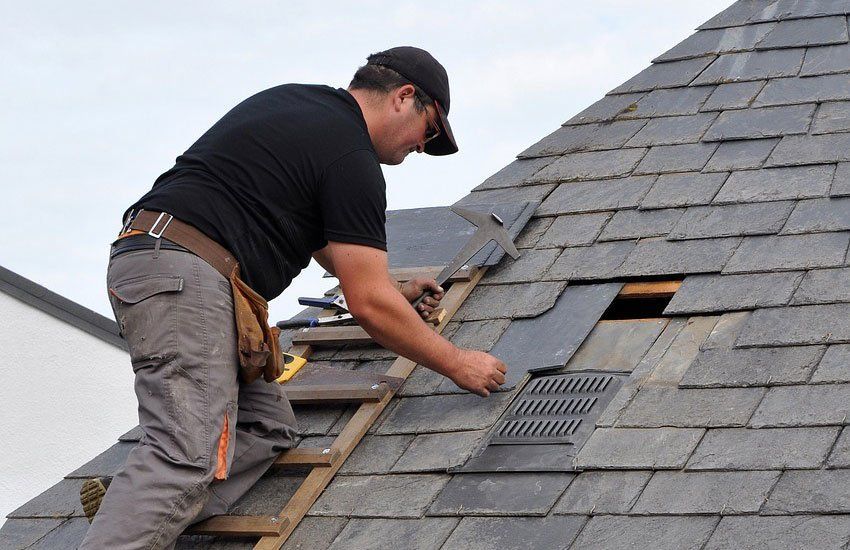 Roofer in black shirt and cap on roof installing slate tiles, near vent.