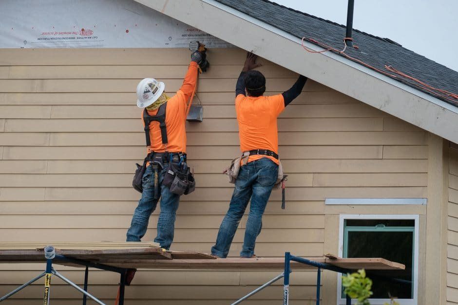 Two construction workers installing siding on a house, working from a scaffold.