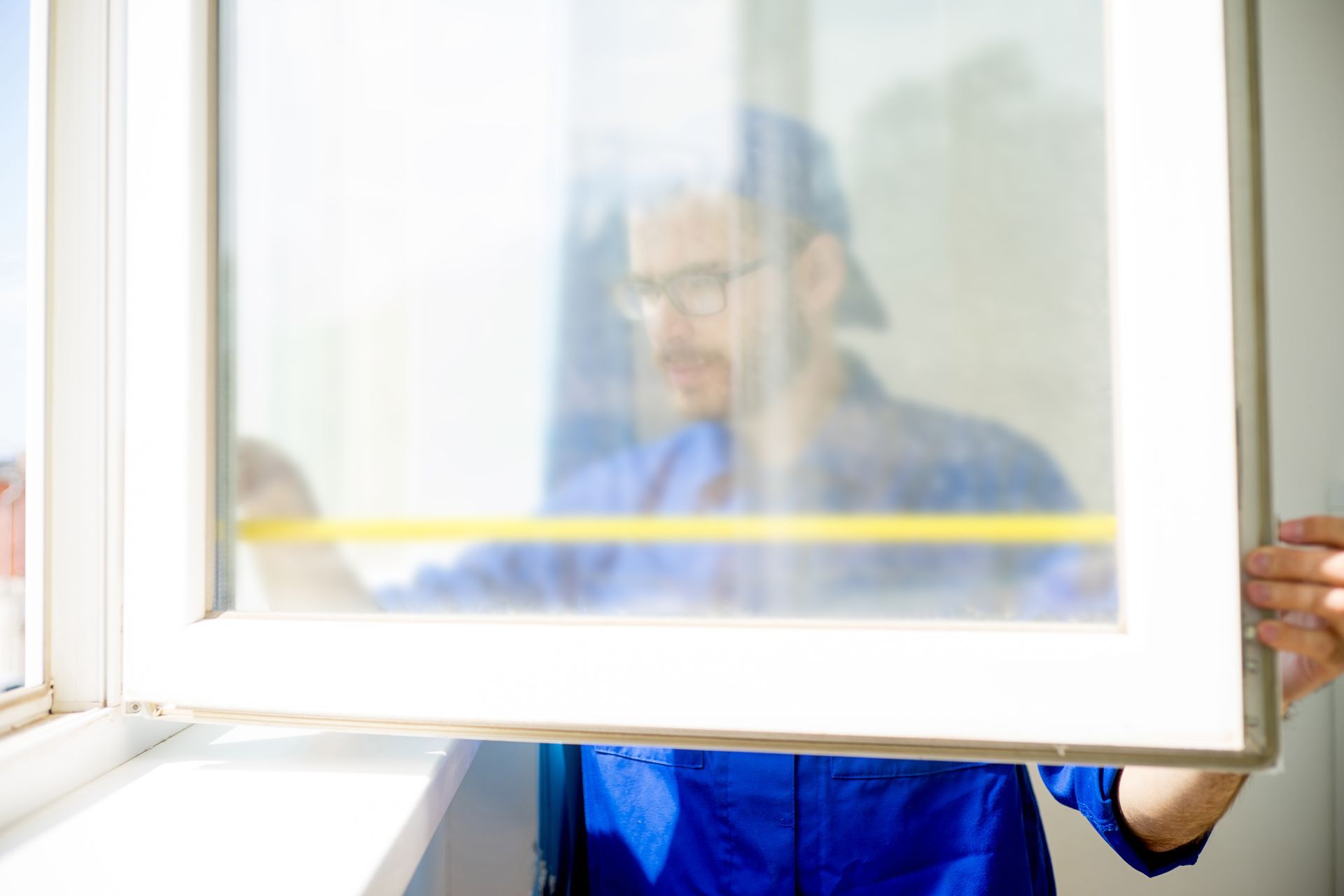Worker measuring and installing a window frame with a partially open glass panel indoors.