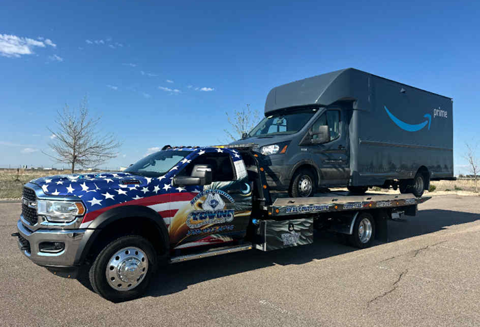 Tow truck with American flag graphics carrying an Amazon Prime delivery van on a sunny day.