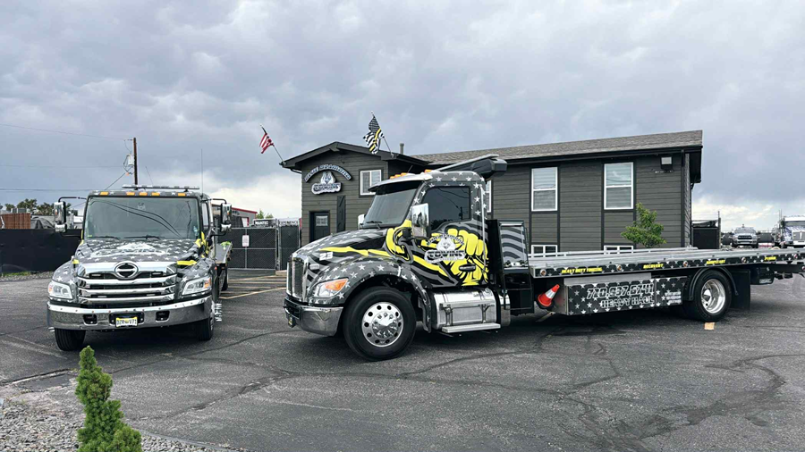 Two tow trucks with yellow and black graphics in front of a building on a cloudy day.