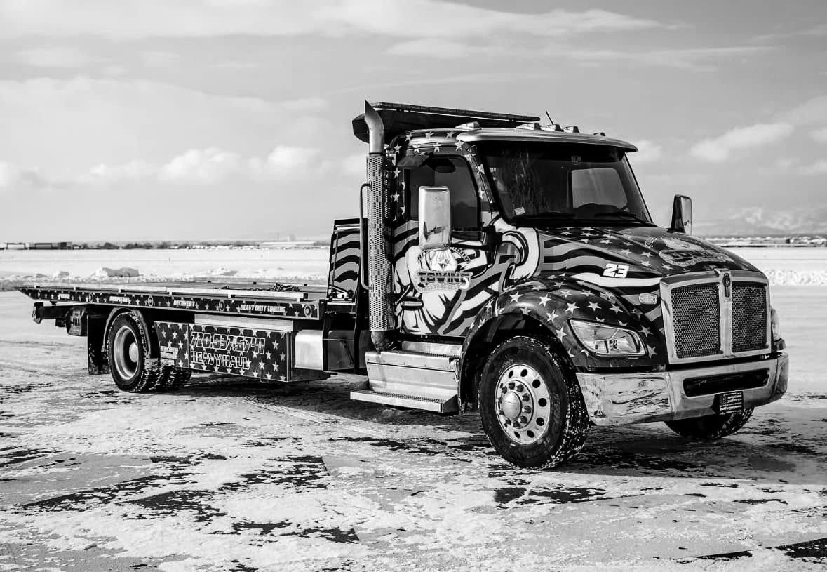 Black and white flatbed tow truck with patriotic paint, parked on a snowy plain.