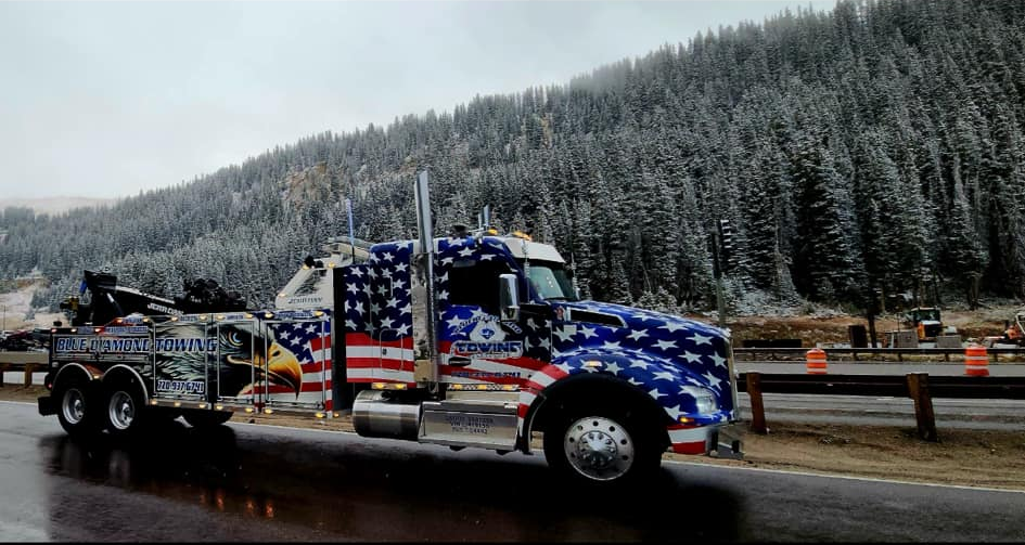 Tow truck with American flag design parked near snow-covered trees.