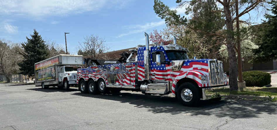 Tow truck decorated with American flag design pulling a smaller truck with a trailer. Parked outdoors.