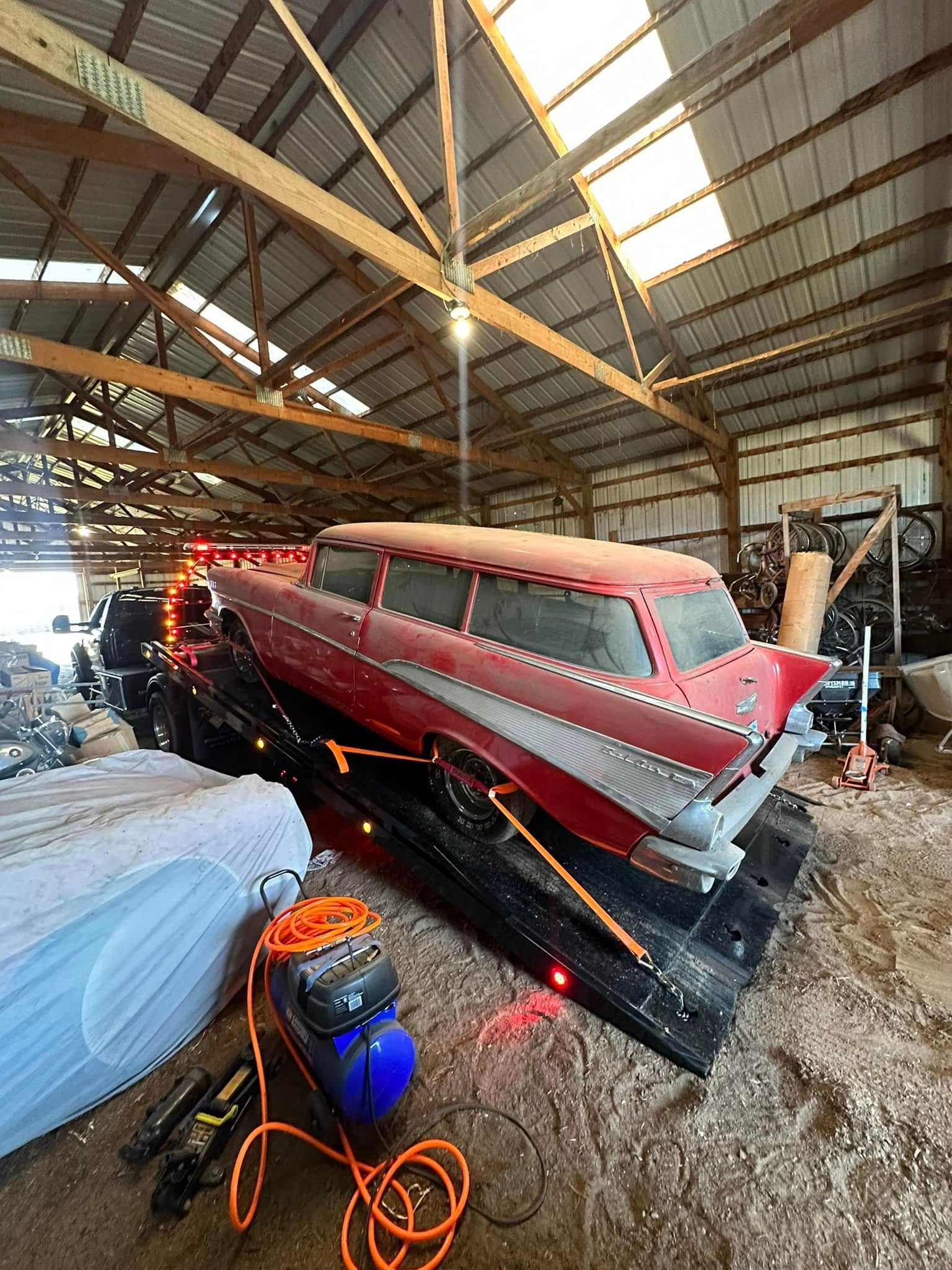 Red classic station wagon on a black trailer inside a wooden barn, with tools and lighting.