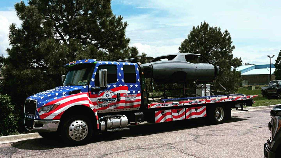 Tow truck with American flag design carrying a large, dark, aircraft part. Outdoors.