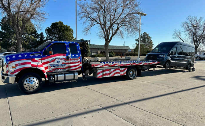 Tow truck with American flag design towing a black van, parked outdoors on a sunny day.