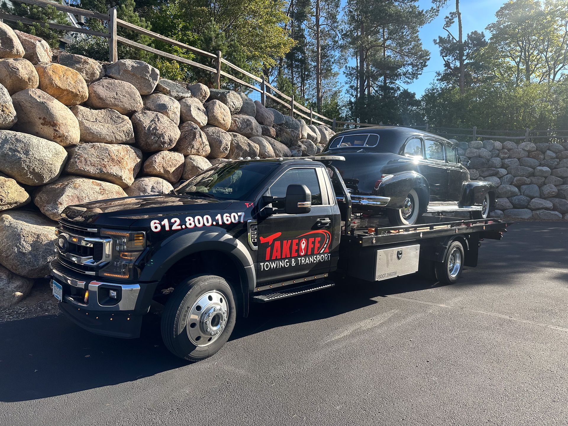 Black tow truck with a classic car on its flatbed, parked in front of a rock wall.