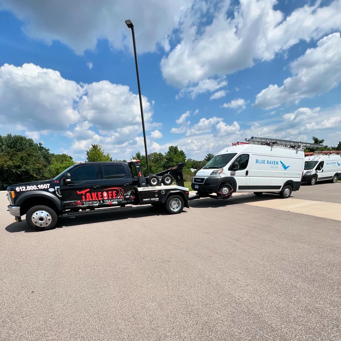 A black tow truck towing a white van, both on a paved surface under a blue sky with clouds.