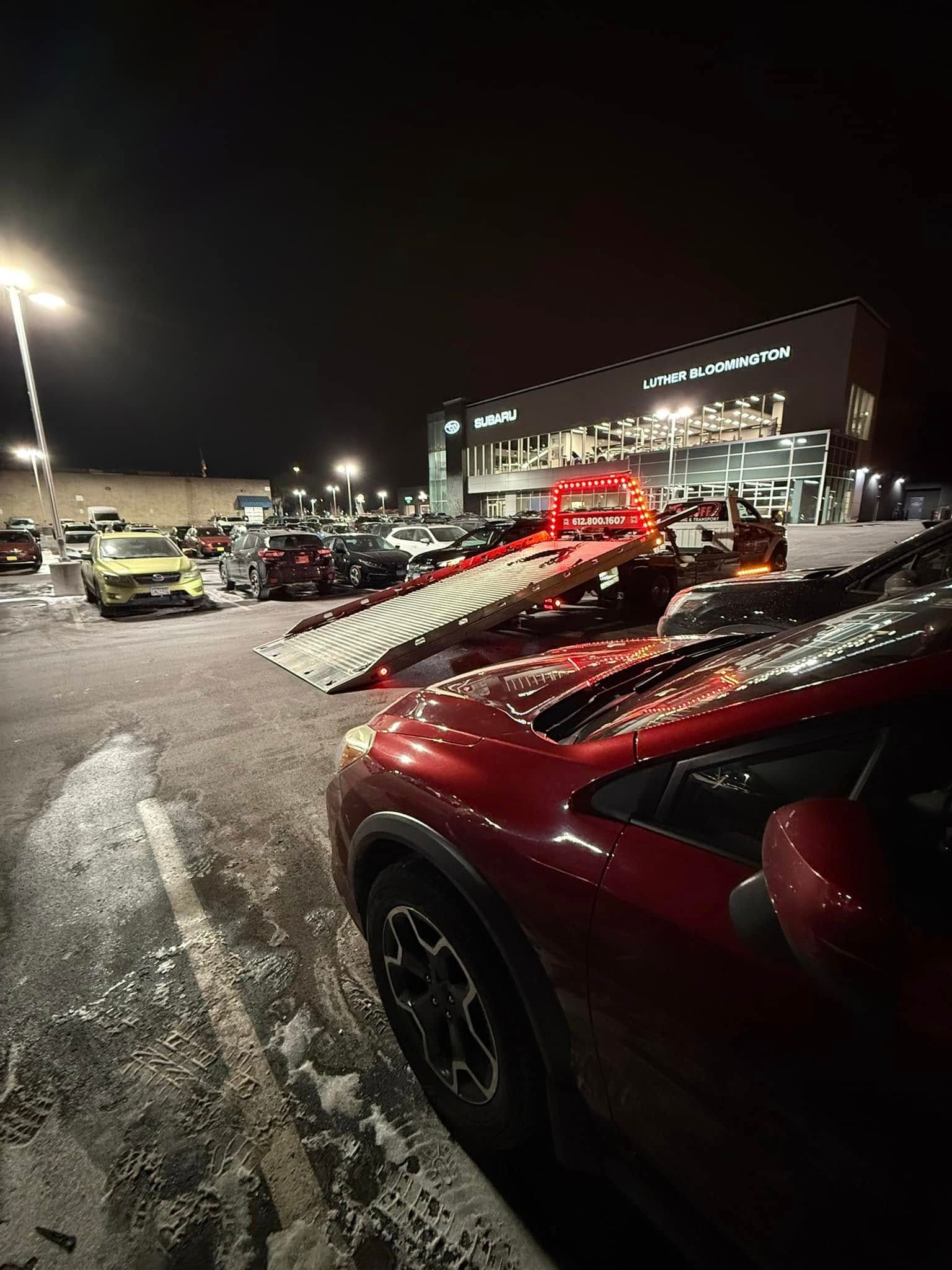 Red SUV being towed at a car dealership at night; snowy lot, building in background.