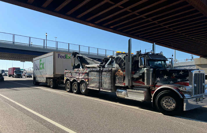 Tow truck towing a FedEx trailer under a bridge on a sunny day. Truck has patriotic stars and stripes design.