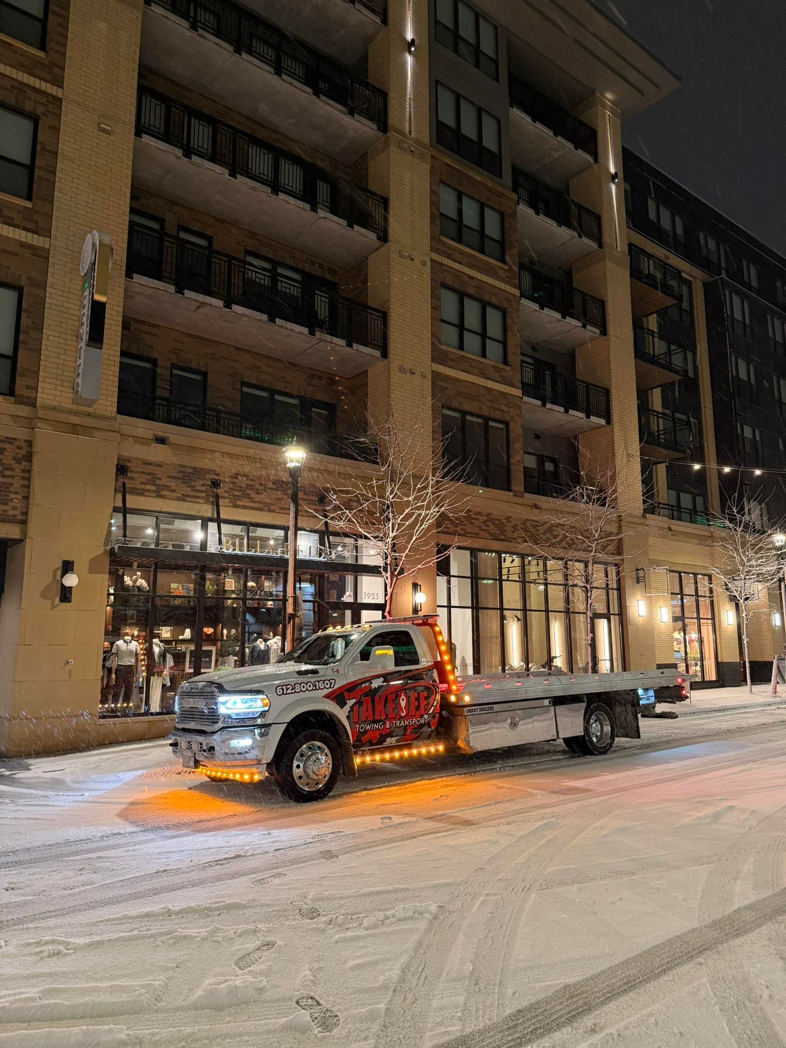 Tow truck in snowy street, lit up, in front of a multi-story building at night.