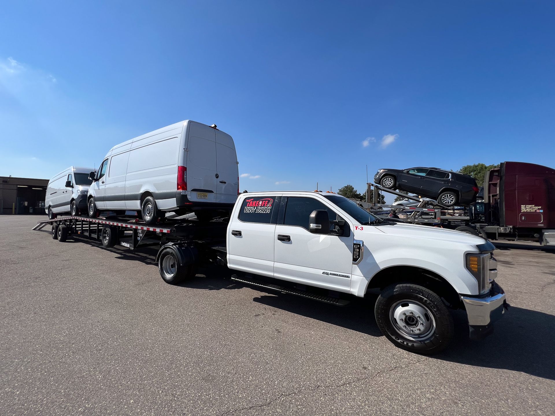 White truck towing a flatbed trailer carrying two vans. A dark-colored car is also on the trailer. Blue sky.
