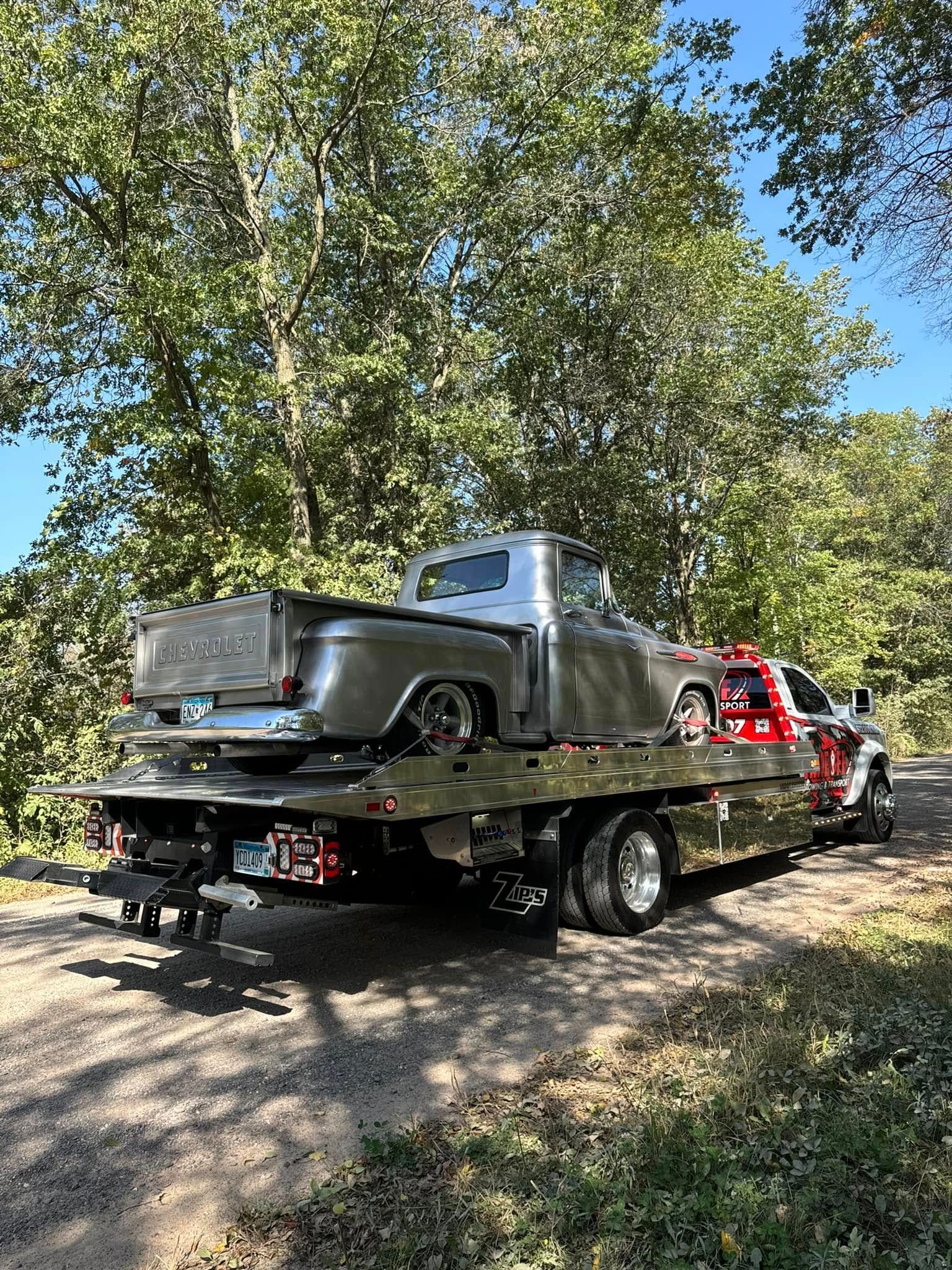 Silver classic truck on a flatbed tow truck, outdoors. Green trees in background, sunny day.
