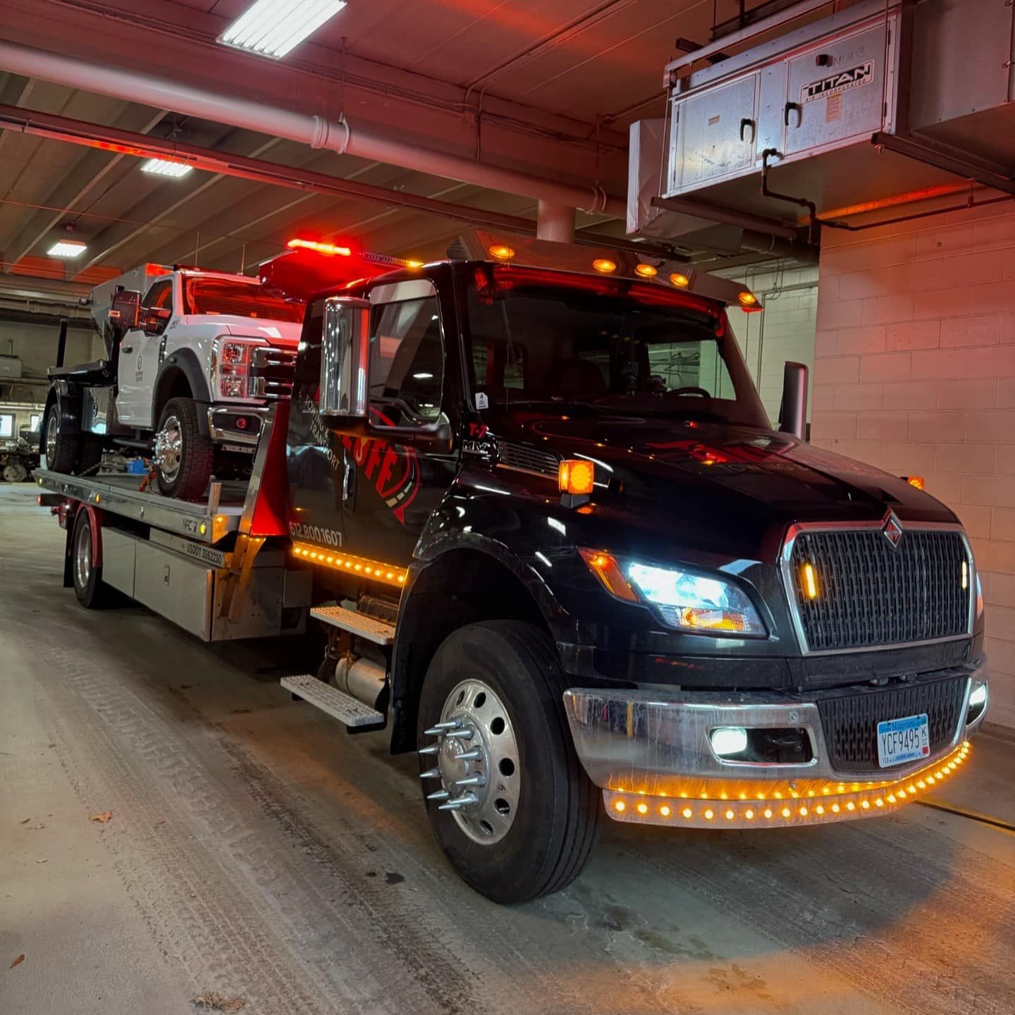 Black tow truck carrying a white truck inside a garage with orange and white lighting.