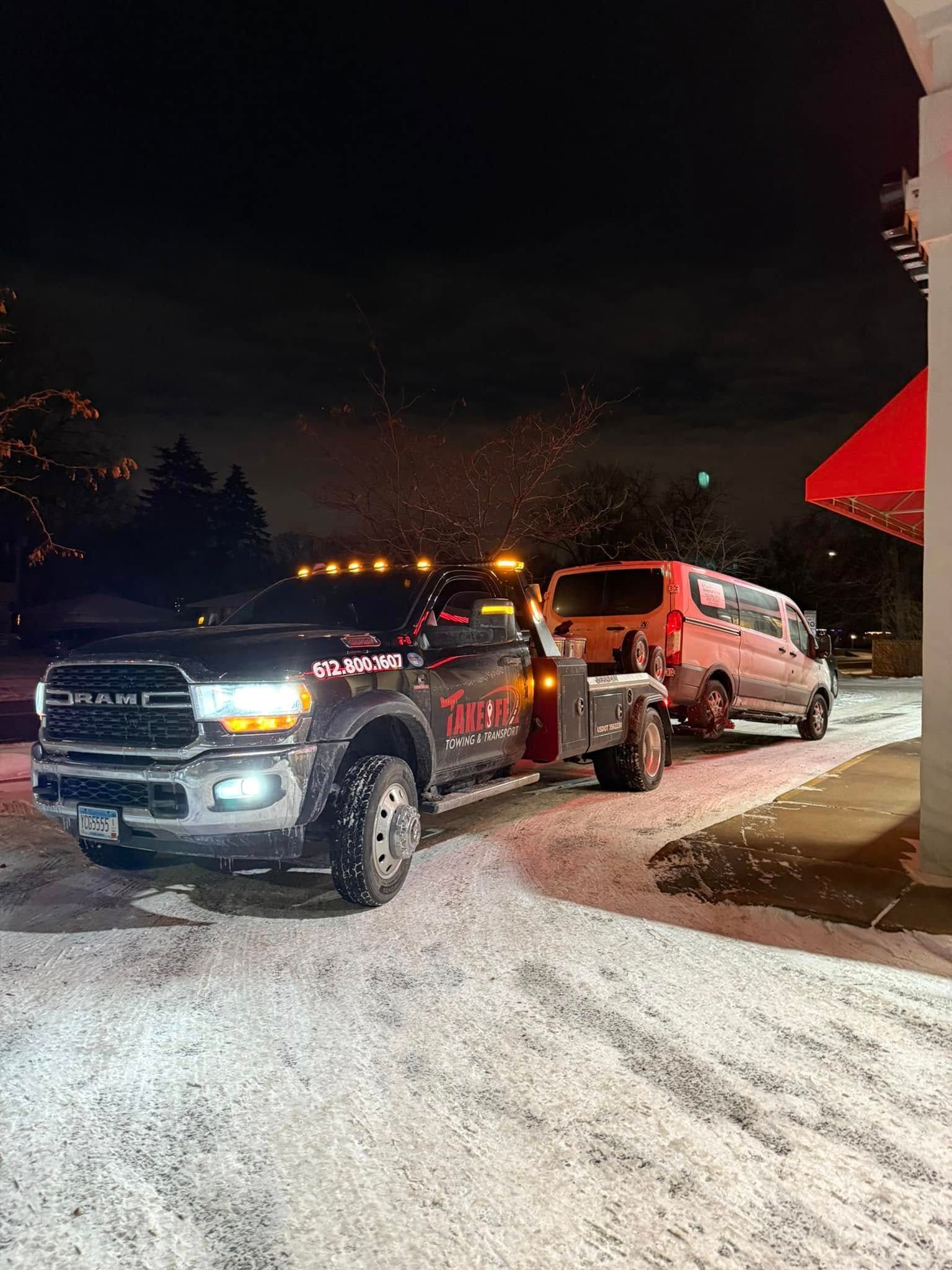 Tow truck towing a van at night in snowy conditions. Red and black truck, white snow.