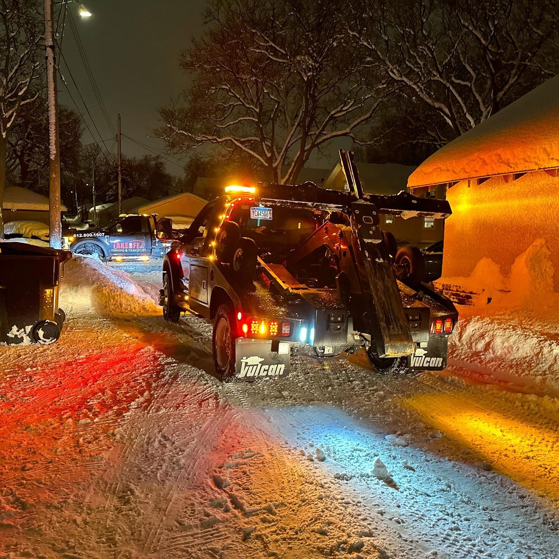 Tow truck on snow-covered street at night, lights flashing.