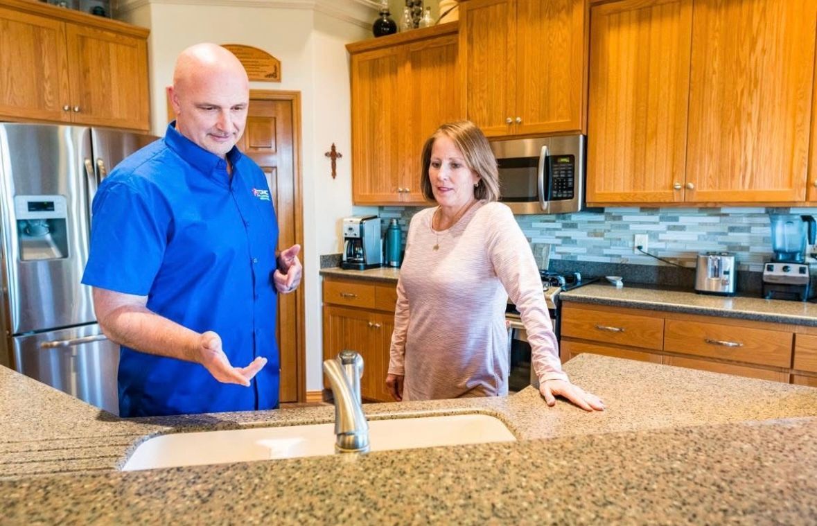 Plumber in blue shirt explains to a woman in a kitchen, sink in foreground.
